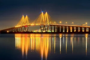 Illuminated suspension bridge reflecting golden lights on calm water at night, showcasing the serene beauty of Baytown, Texas.