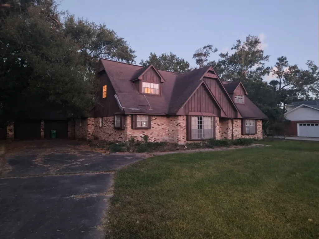 A brick and wood two-story home with a sloping roof and garage, nestled among trees with a large front lawn at dusk.